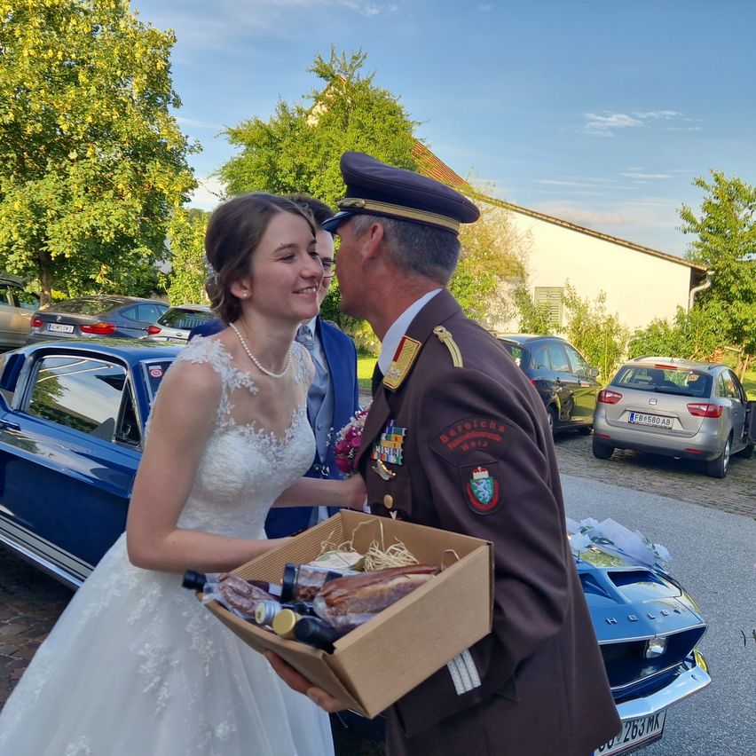 A bride in a white gown holds a box of food near a military officer in uniform, with cars and trees in the background.