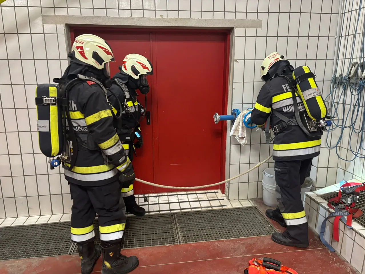 Drei Feuerwehrleute in voller Ausrüstung bereiten sich darauf vor, eine rote Tür zu betreten, einer hält einen Schlauch und ein anderer ein Tuch.