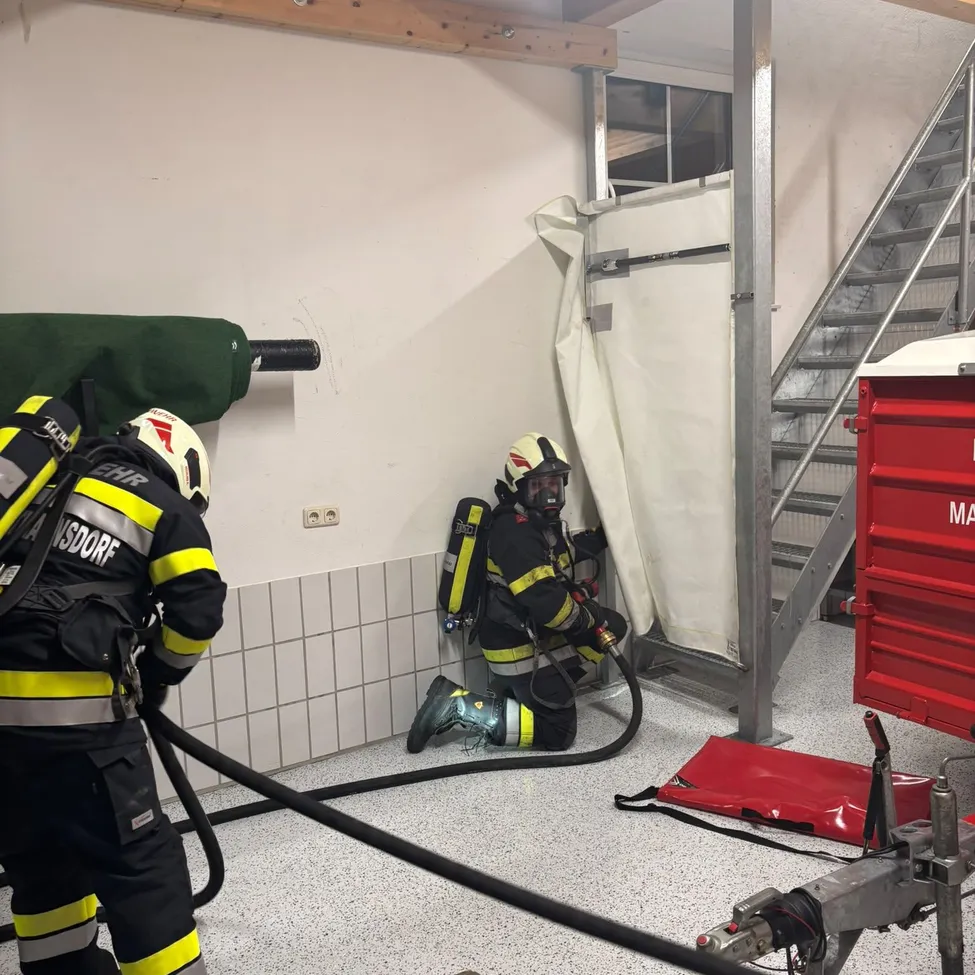 Zwei Feuerwehrleute in voller Ausrüstung üben in einem Raum mit einem roten Container und Treppen.