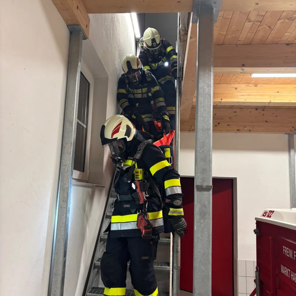 Drei Feuerwehrleute in voller Ausrüstung steigen eine Treppe hinauf, einer hält ein rotes Objekt. Die Umgebung ist innen, mit einer Holzdecke und weißen Wänden.