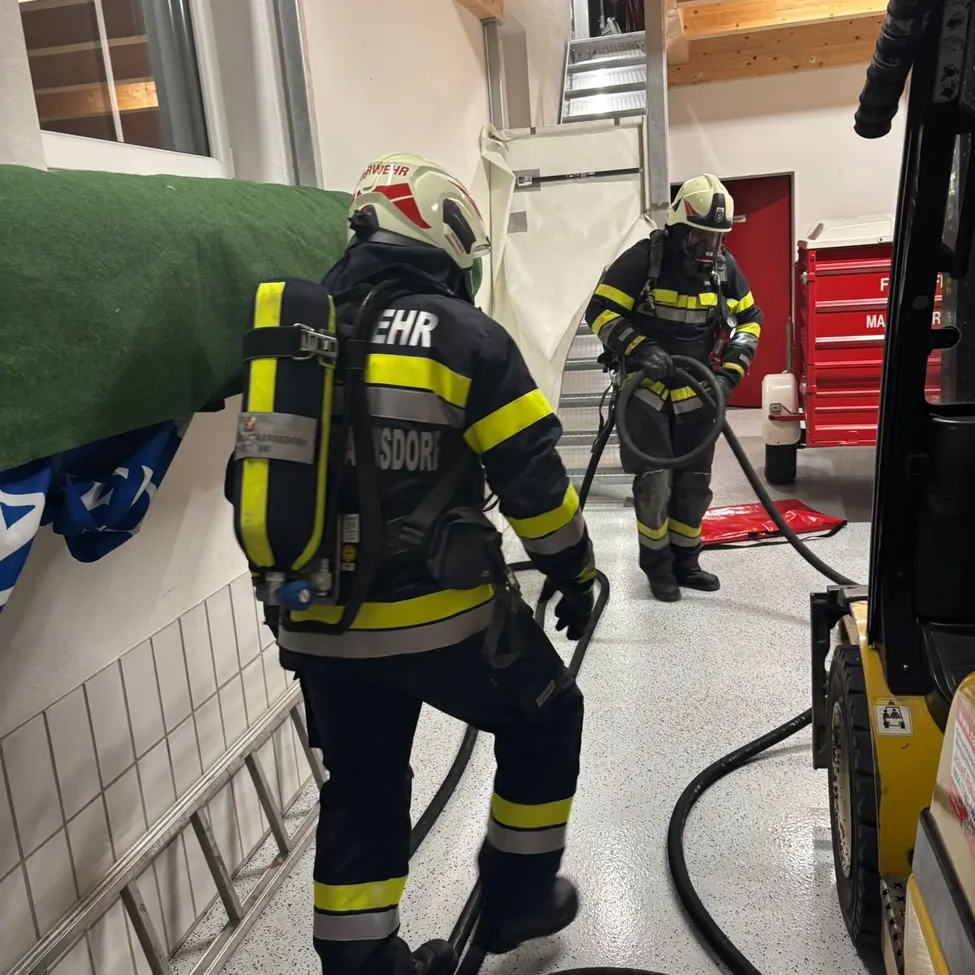 Zwei Feuerwehrleute in voller Ausrüstung arbeiten in einem Raum, einer hält einen Schlauch und der andere trägt Ausrüstung. Ein rotes Fahrzeug ist in der Nähe.