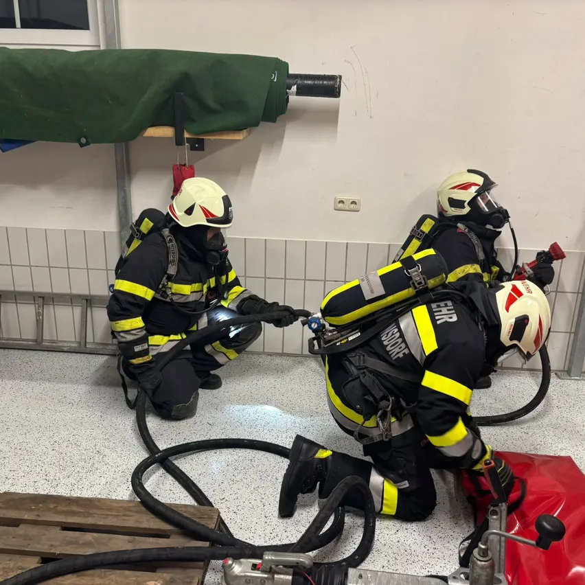 Drei Feuerwehrleute in voller Ausrüstung, einer kniet und hält einen Schlauch, ein anderer hält einen roten Beutel und der dritte steht mit einem Werkzeug. Sie befinden sich in einem Raum mit weißen gefliesten Wänden und einem Holzpalette.