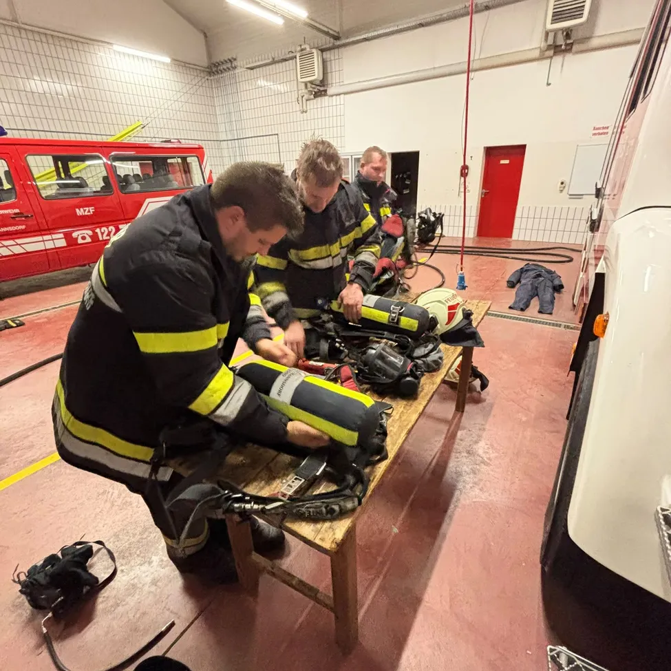 Drei Feuerwehrleute arbeiten an einem Holztisch, umgeben von Ausrüstung und einem Feuerwehrwagen im Hintergrund.