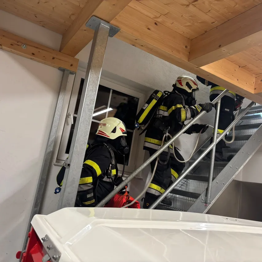 Feuerwehrleute in voller Ausrüstung steigen eine Treppe hinauf, einer hält ein Seil. Sie sind mit Helmen und Masken ausgestattet. Die Szene spielt drinnen, mit einer Holzdecke und einem Fenster.