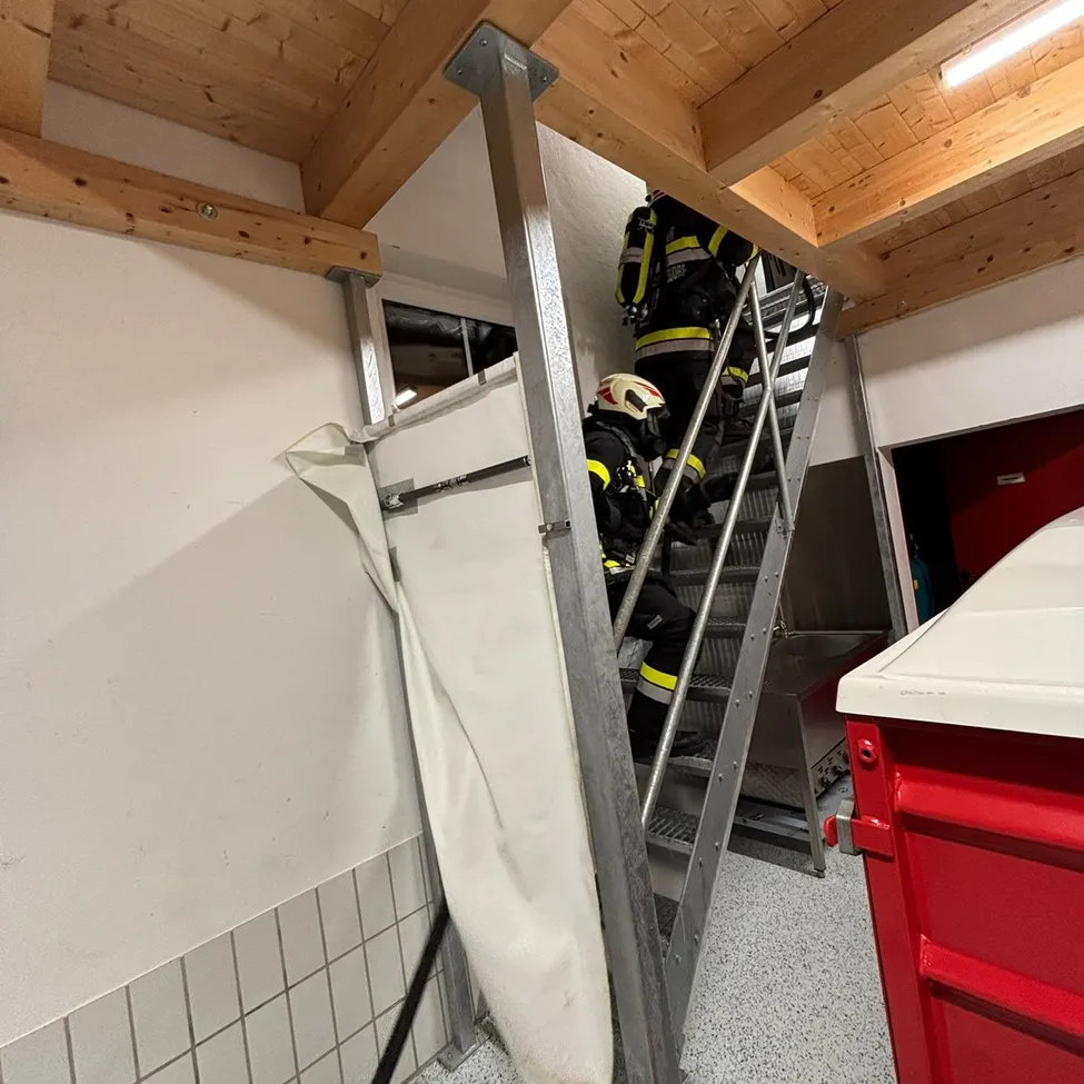 Feuerwehrleute klettern eine Metallleiter in einem Gebäude. Einer trägt einen Helm und eine reflektierende Weste. Der Raum hat eine Holzdecke und einen gefliesten Boden.