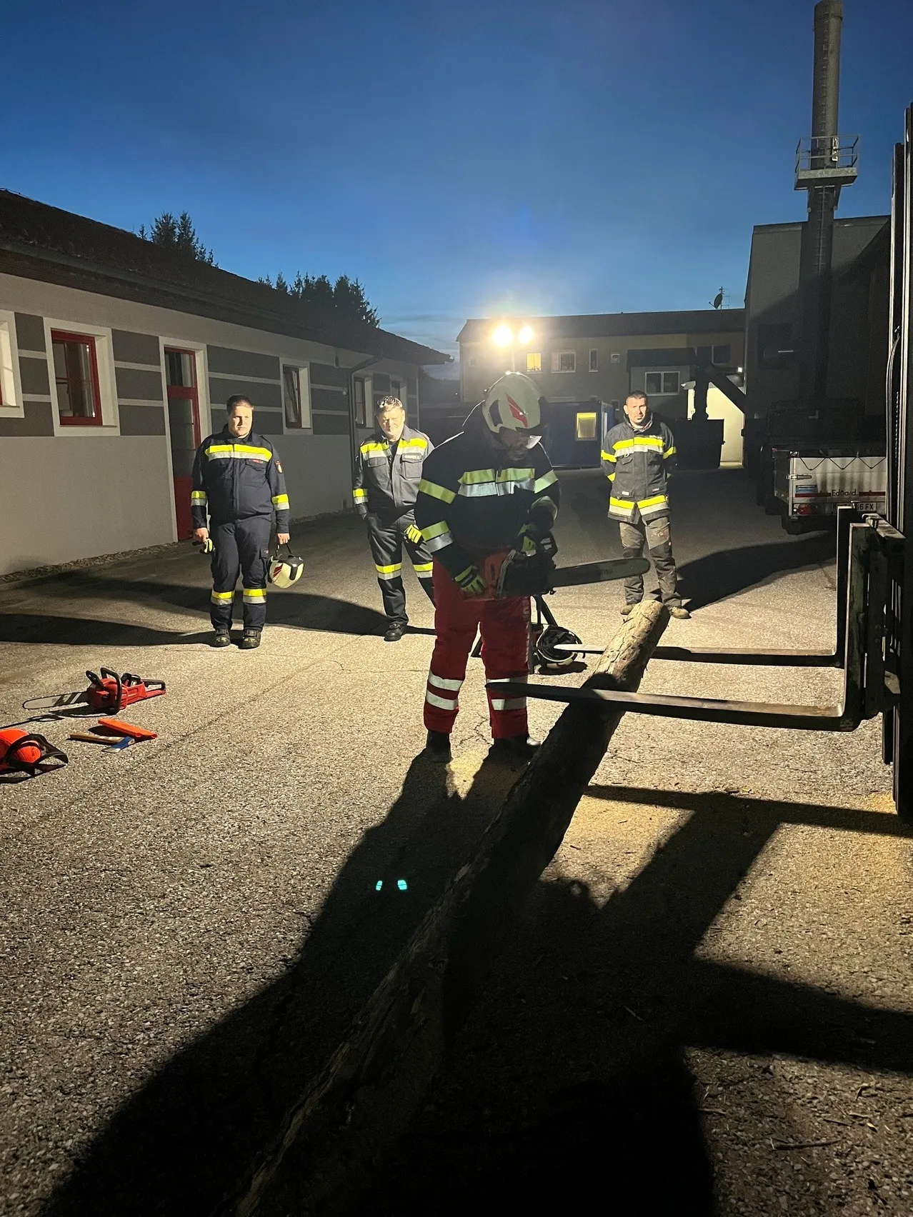Vier Feuerwehrleute stehen nachts auf einem Parkplatz, einer von ihnen hält eine Kettensäge und schneidet einen Baumstamm.