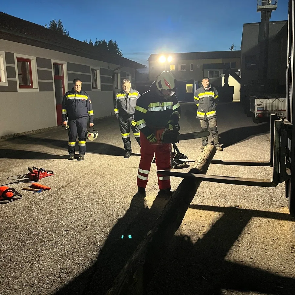 Vier Feuerwehrleute stehen nachts auf einem Parkplatz, einer von ihnen hält eine Kettensäge und schneidet einen Baumstamm.
