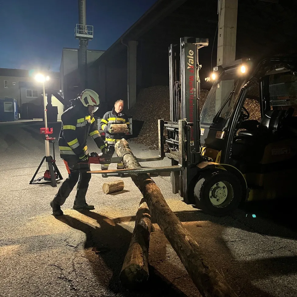 Zwei Feuerwehrleute schneiden mit einer Kettensäge Holz unter der Anleitung eines Gabelstaplers bei Nacht.