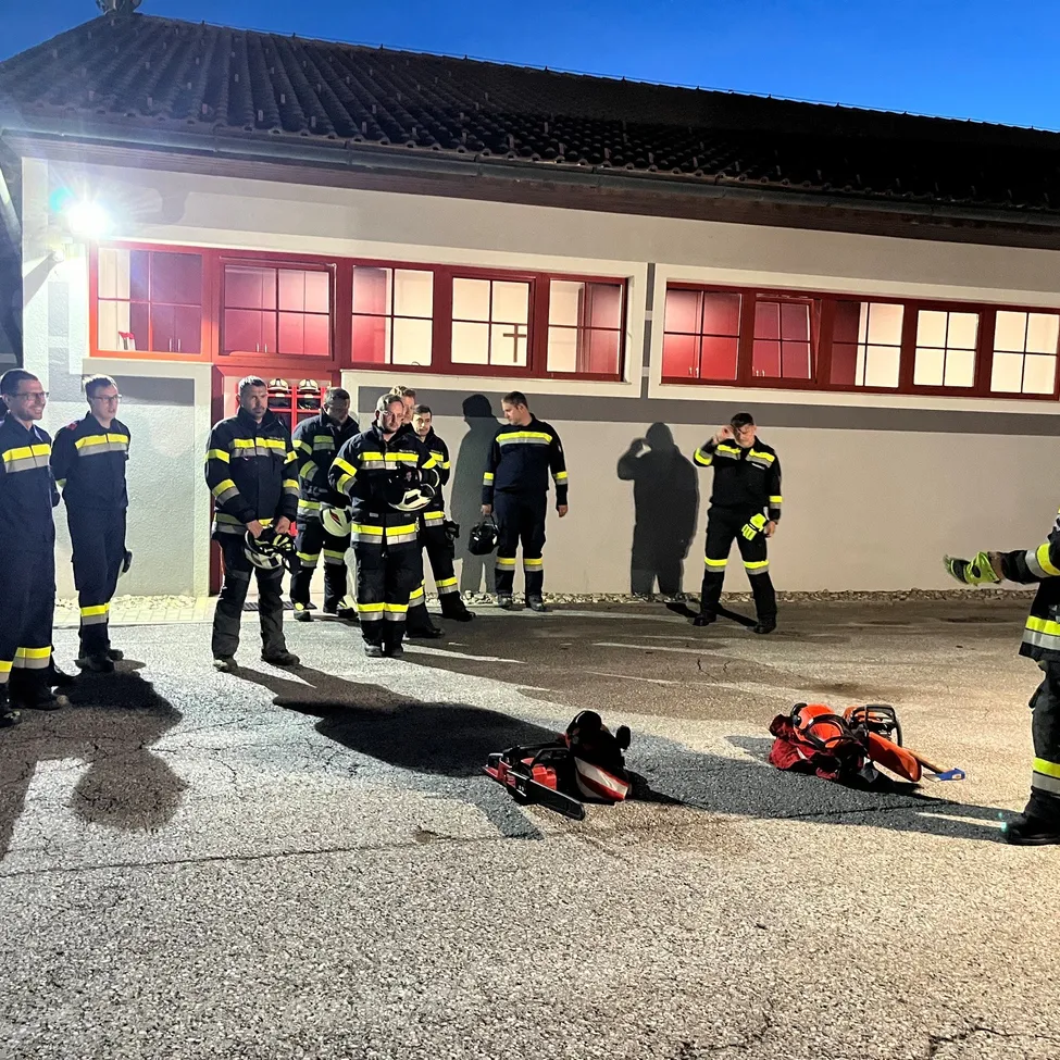 Eine Gruppe von Feuerwehrleuten ist nachts vor einer Feuerwache versammelt, wobei eine Person einen Helm hält und eine andere Person ein Werkzeug in der Hand hat.