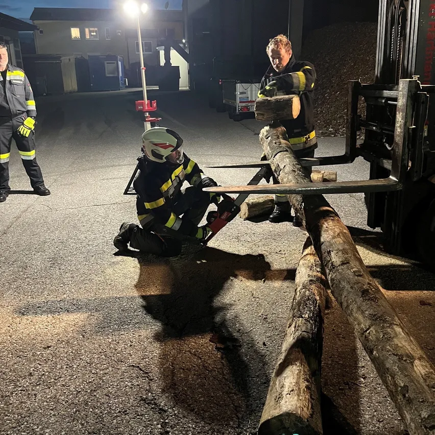 Zwei Feuerwehrleute arbeiten mit einer Kettensäge an einem Baumstamm, einer kniet und der andere steht hinter ihm.