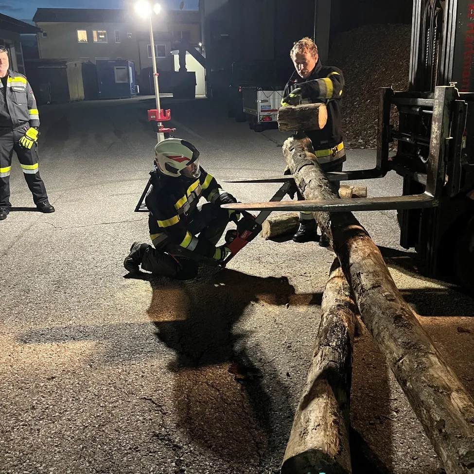 Zwei Feuerwehrleute arbeiten mit einer Kettensäge an einem Baumstamm, einer kniet und der andere steht hinter ihm.