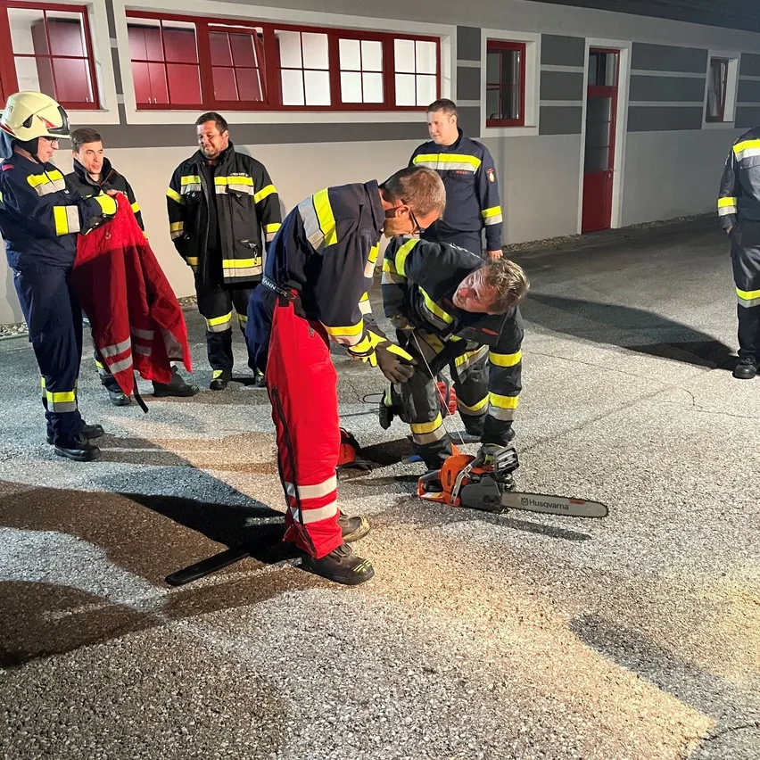 Eine Gruppe von Feuerwehrleuten in Uniform versammelt sich um einen Mann, der einen Kettensägen auf dem Boden benutzt.