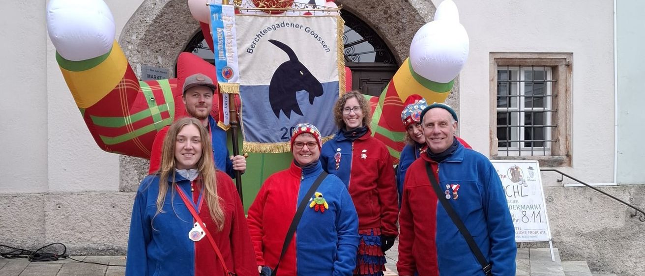 Eine Gruppe von Menschen in farbenfrohen Kostümen posiert vor einer riesigen Ballonfigur eines Clowns. Der Clown hält ein Banner mit den Worten 'Bergensgadener Goaßen'. Die Menschen stehen auf Stufen vor einem Gebäude.