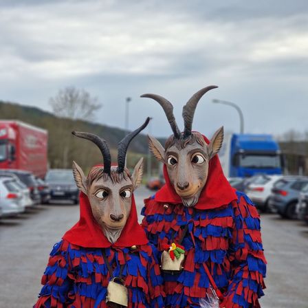 Zwei Personen in Ziegenmasken und roten Kapuzen stehen auf einem Parkplatz, umsäumt von Lastwagen und Autos.