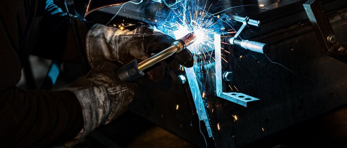 Eine Person mit Handschuhen schweißt ein Metallstück mit einem Schweißbrenner, wobei Funken entstehen. Der Schweißer trägt eine Maske zum Schutz.