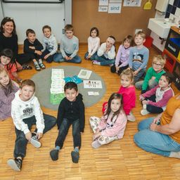 Eine Gruppe von Kindern sitzt auf dem Boden in einem Klassenzimmer. Sie sitzen im Kreis um einen Tisch mit Büchern und Papieren. Die Lehrerin sitzt auf der linken Seite der Kinder.