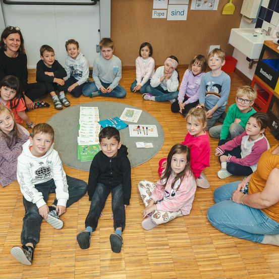 Eine Gruppe von Kindern sitzt auf dem Boden in einem Klassenzimmer. Sie sitzen im Kreis um einen Tisch mit Büchern und Papieren. Die Lehrerin sitzt auf der linken Seite der Kinder.