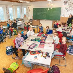 Ein Klassenzimmer mit Kindern an Schreibtischen, ein Lehrer steht in der Nähe der Tafel, und ein Klassenzimmer gefüllt mit verschiedenen Schulsachen und Rucksäcken.