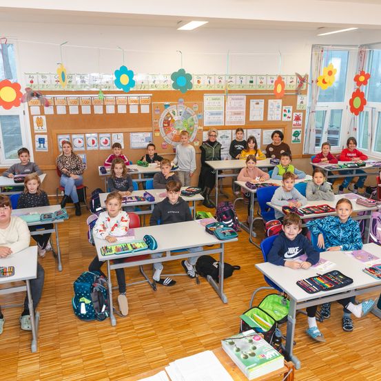 Klassenzimmerszene mit Schülern an Schreibtischen, farbenfrohen Blumen am Bulletin Board und einer Lehrerin vorne stehend.