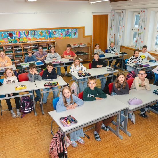 Ein Klassenzimmer voller Schüler an Schreibtischen, einige schauen in die Kamera. Der Raum hat Holzböden, weiße Wände und bunte Kunstwerke. Rucksäcke und Bücher sind auf den Schreibtischen.