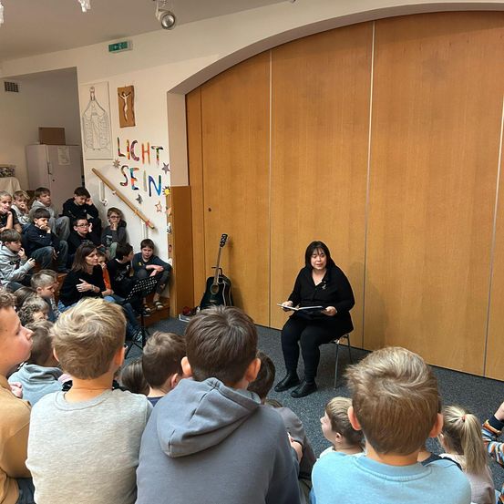 Eine Frau sitzt auf einem Stuhl vor einer Wand und liest ein Buch vor einer Gruppe von Kindern, die aufmerksam zuhören. Die Wand hat ein Gemälde und die Worte 'Licht' und 'Sein'.