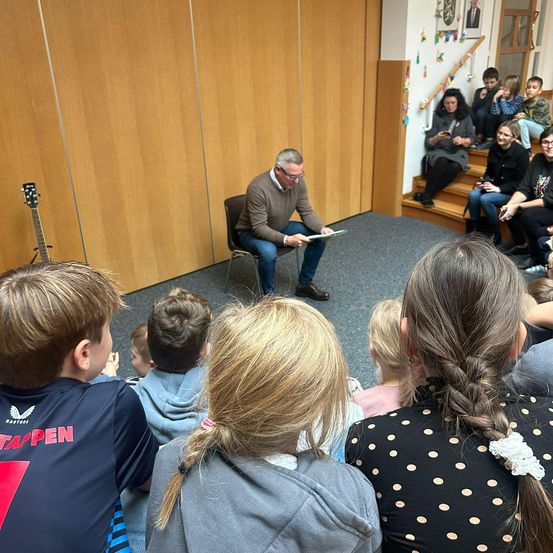 Ein Mann liest ein Buch vor einer Gruppe von Kindern, die auf dem Boden in einem Raum sitzen. Hinter ihm steht eine Gitarre. Der Raum hat Holzwände und eine Treppe.