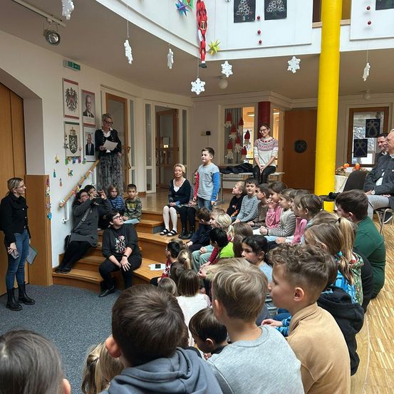 Eine Gruppe von Kindern und Erwachsenen versammelt sich in einem Raum mit Holzboden. Zwei Frauen stehen auf einer Plattform und halten möglicherweise eine Rede. Der Raum ist mit Dekorationen geschmückt, die von der Decke hängen.