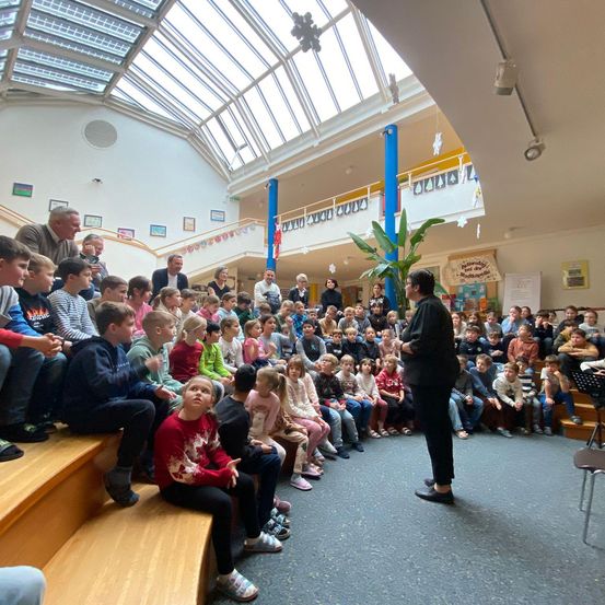 Eine große Gruppe von Kindern sitzt auf Bänken und auf dem Boden in einem Raum. Ein Mann steht vor ihnen, und ein Topf mit Pflanzen steht neben ihm. Es gibt ein Geländer auf der oberen Etage, und einige Dekorationen hängen von der Decke.
