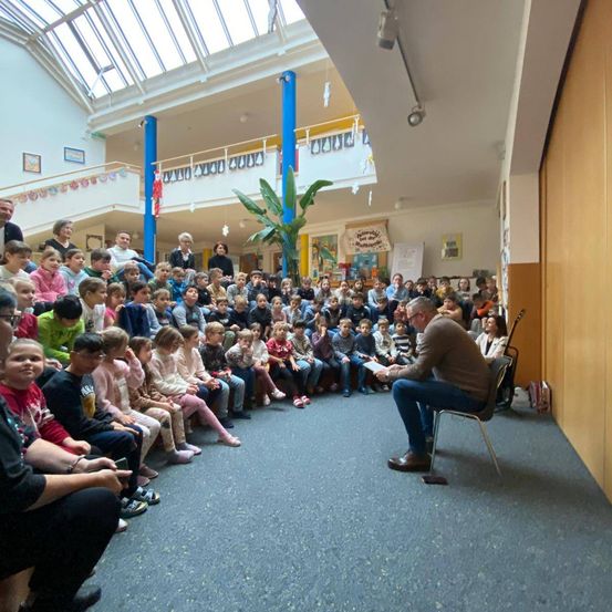 Ein Mann liest einer großen Gruppe von Kindern in einem Raum mit Glasdecke und weißen Wänden vor.