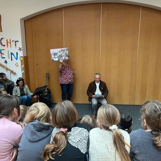 Ein älterer Mann liest ein Buch im Sitzen, während eine Frau mit einem Foto dahinter steht. Ein Publikum von Kindern sitzt davor und eine Gitarre steht auf einem Notenständer.