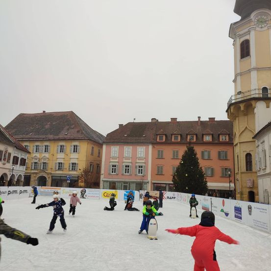 Mehrere Menschen eislaufen auf einer Eisbahn in einem Stadtplatz, mit Gebäuden und einem Weihnachtsbaum im Hintergrund.
