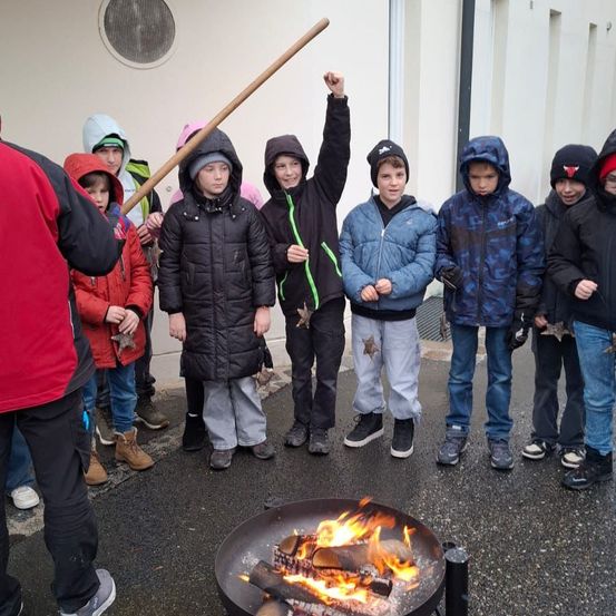 Eine Gruppe von Kindern steht um ein Feuer herum und hält Marshmallow-Sticks. Sie tragen Winterkleidung, und ein Junge hebt die Hand. Das Feuer brennt in einem Metallbecken.
