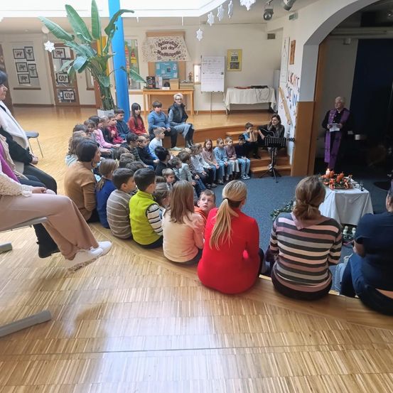 In einem Kirchensaal sitzen eine Gruppe von Kindern und Erwachsenen um eine Bühne herum, auf der eine Frau Gitarre spielt und ein Mann liest. Ein Tisch mit Blumen und Kerzen steht vorne.