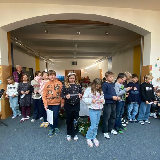 Eine Gruppe von Kindern steht in einem Raum, hält Papiere in den Händen und scheint an einer Veranstaltung oder Zeremonie teilzunehmen. Ein Mann steht hinter ihnen, und einige Dekorationen sind an der Wand angebracht.
