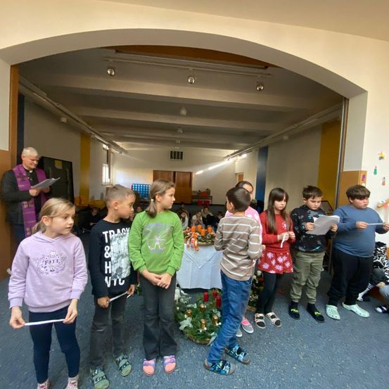 Kinder stehen in einem Raum und halten Papiere in der Hand, möglicherweise singen sie. Ein Mann in einer violetten Stola steht vor ihnen. Ein Tisch mit Kerzen steht hinter ihnen und eine Wand mit Dekorationen befindet sich rechts.