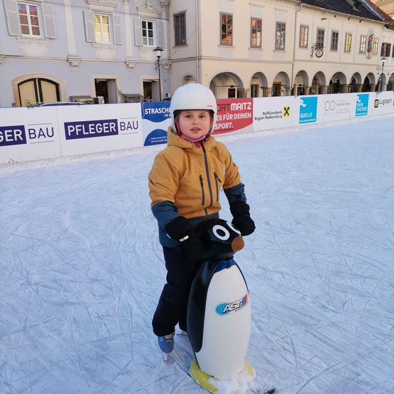 Ein junges Kind in einer gelben Jacke fährt auf einem pinguinförmigen Schlitten auf einer Eisbahn, mit einem Gebäude mit Bogenfenstern im Hintergrund.