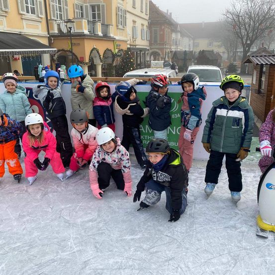 Eine Gruppe von Kindern in Winterkleidung und Helmen posiert für ein Foto auf einer Eisbahn mit einem Pinguin-Standbild im Hintergrund.