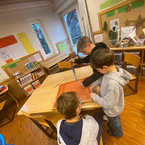 Drei Jungen arbeiten in einem Klassenzimmer mit Holzböden an einem Projekt. Sie konzentrieren sich auf ein orangefarbenes Stück Stoff. Der Raum hat Tische, Stühle und Bücherregale.