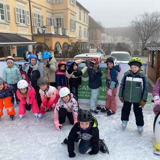 Eine Gruppe von Kindern in Winterkleidung und Helmen eislaufen auf einer Eisbahn mit einem Banner dahinter.