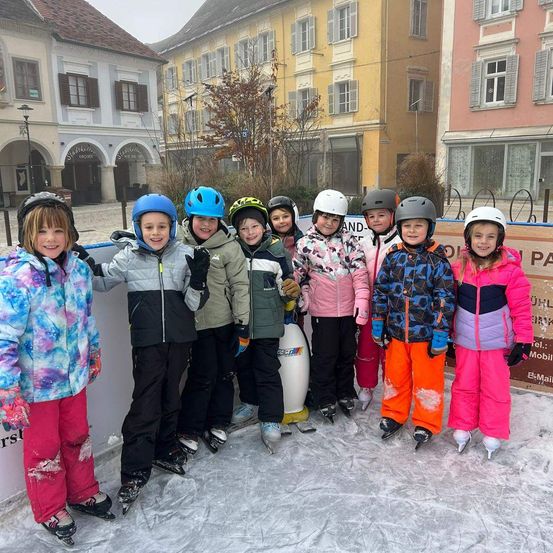 Eine Gruppe von Kindern mit Helmen und Winterkleidung steht zusammen auf einer Eisbahn vor einem Gebäude.