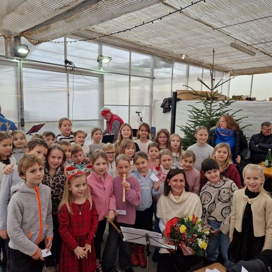 Eine Gruppe von Kindern und Erwachsenen befindet sich in einem Gewächshaus. Die Kinder halten Musikinstrumente und einige tragen Mützen. Eine Frau vorne hält einen Blumenstrauß. Ein Weihnachtsbaum ist im Hintergrund.