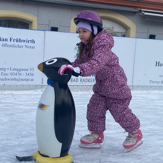 Ein kleines Mädchen mit Helm und Schlittschuhen hält eine Pinguinstatue fest, während es auf einer Eisbahn Schlittschuh läuft. Hinter ihr steht ein Gebäude mit einem Schild, das 'Bad Radkersburg' besagt.