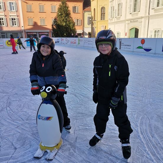 Zwei Kinder stehen auf einer pinguinförmigen Skimaschine auf einer Eisbahn und lächeln für die Kamera. Im Hintergrund befinden sich Gebäude mit Weihnachtsbäumen und Menschen, die skaten.