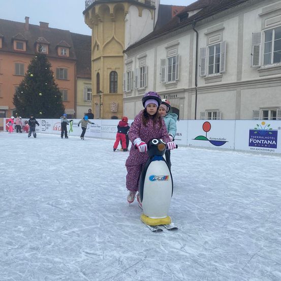 Ein Mädchen fährt auf einem pinguinförmigen Eisschlitten auf einer Eislaufbahn im Freien, mit Gebäuden und einem Weihnachtsbaum im Hintergrund.