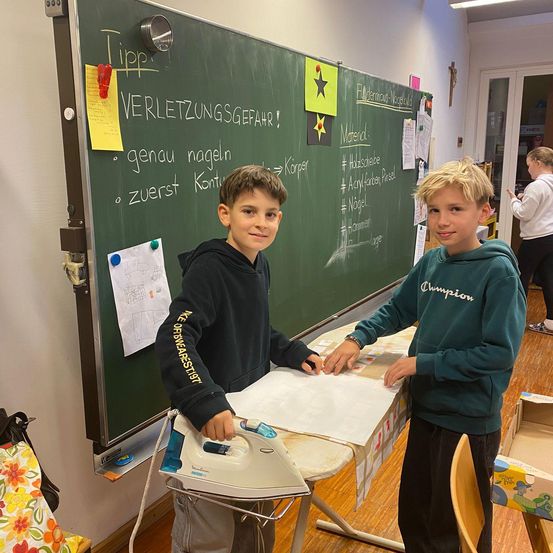 Zwei Jungen stehen vor einem Tisch und benutzen ein Bügeleisen. Sie lächeln und scheinen an einem Projekt zu arbeiten. Dahinter ist eine Tafel mit deutschem Text zu sehen. Andere Gegenstände und Personen sind im Raum.