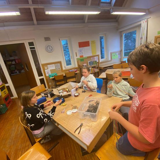 Vier Kinder arbeiten an einem Projekt an einem Tisch im Klassenzimmer. Sie sind von verschiedenen Werkzeugen und Materialien umgeben. Der Raum hat große Fenster und eine Uhr an der Wand.