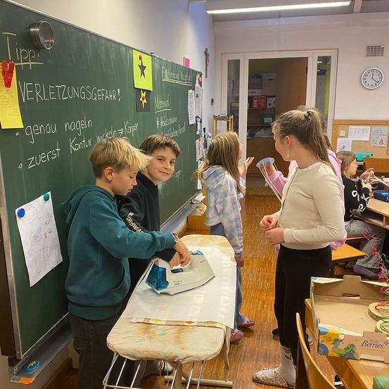 In einem Klassenzimmer stehen vier Kinder um einen Bügeltisch herum. Ein Junge benutzt das Bügeleisen, während ein Mädchen in der Nähe steht. Dahinter beobachtet eine Lehrerin. Die Tafel hat deutschen Text, und es gibt Dekorationen und eine Uhr an der Wand.