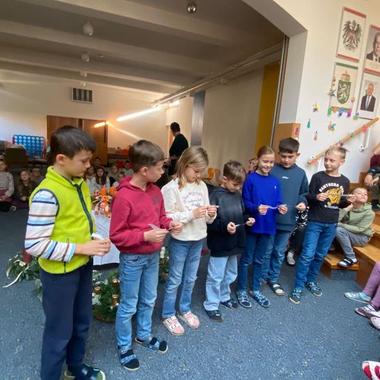 Mehrere Kinder stehen in einer Reihe und halten kleine Kerzen. Sie befinden sich in einem Raum mit weißen Wänden und blauem Teppich. Dahinter sitzen einige Personen auf Treppenstufen.