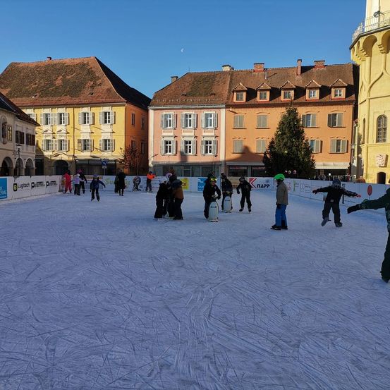 Eine Gruppe von Menschen, die auf einer Freiluft-Eisbahn umgeben von historischen Gebäuden Schlittschuh laufen. Einige tragen Winterkleidung und Helme.