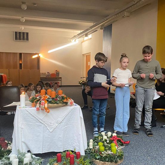 Drei Kinder stehen vor einem dekorierten Tisch mit Kerzen. Sie befinden sich in einem Raum mit hellem Licht, mit anderen Kindern im Hintergrund.